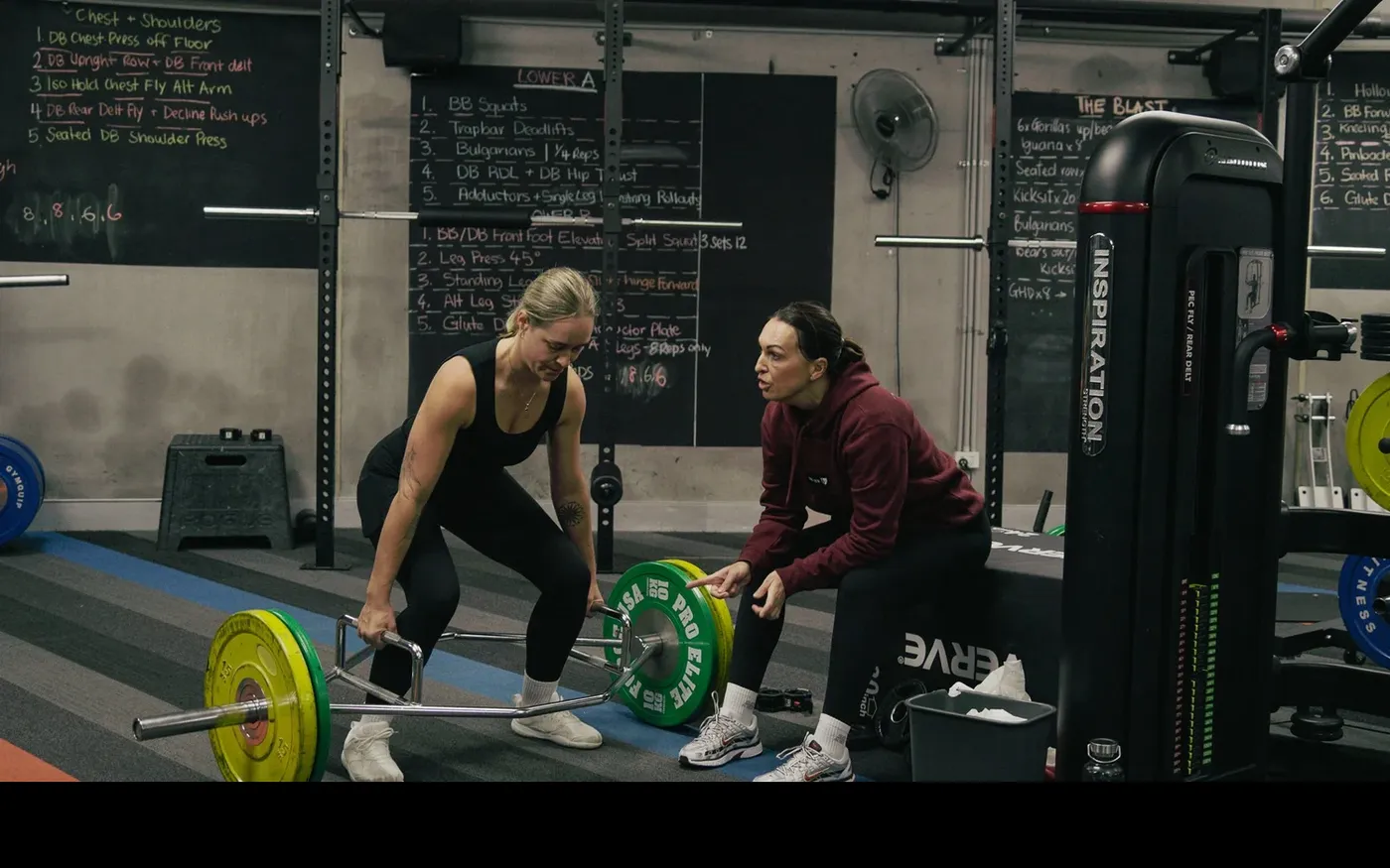 Nicole coaching a member through a trap-bar deadlift in the weights area